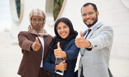 Multicultural business group posing and making like gesture. Businessman and Muslim businesswomen standing outdoors, showing thumb up and smiling at camera. Successful business team concept
