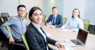 Smiling Business Lady Working with Colleagues Four smiling middle-aged multi-ethnic business people turning to camera, working and discussing ideas while sitting at big table in conference room. Side view.