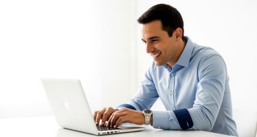 Man Working on Laptop Focused on Task. A man sits at a table, focused on his laptop, smiling slightly.